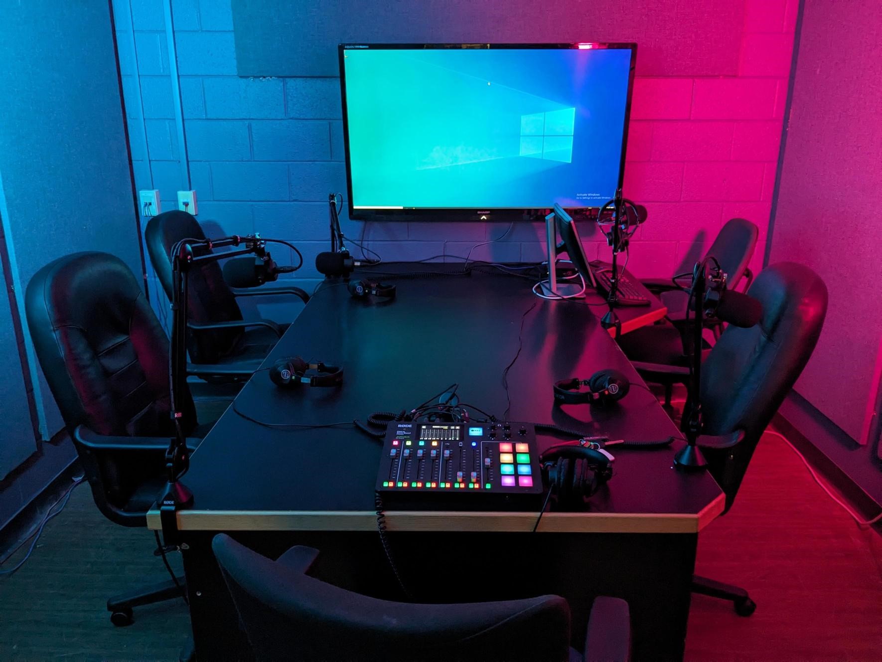 photo of table with 4 chairs, 4 microphones, audio console, video monitor